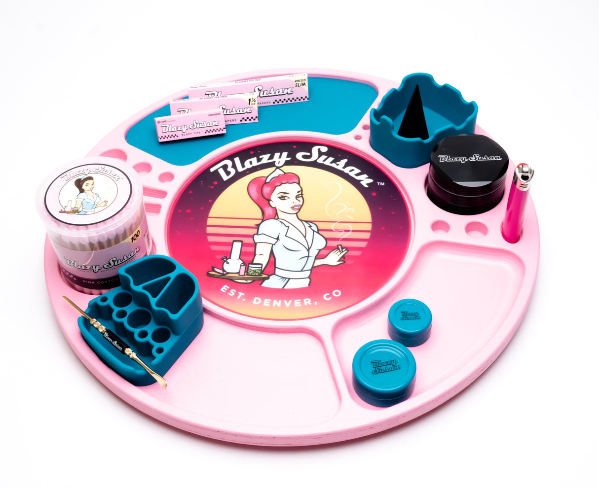Blazy Susan pink spinning rolling tray with compartments, holding branded rolling papers, herb grinder, silicone holders, lighter, dab tool, stash jars, and cotton swabs.