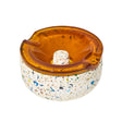 Symi Ashtray featuring a terrazzo-style concrete base with colored chips and an amber-tinted glass insert, showcasing grooves for cigarette rests.