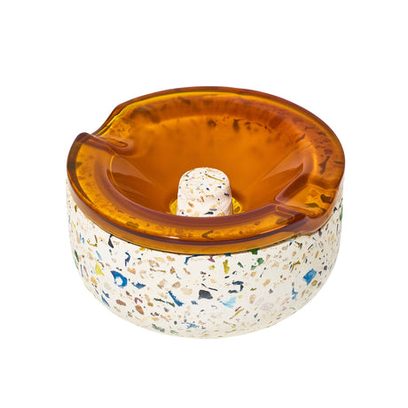 Symi Ashtray featuring a terrazzo-style concrete base with colored chips and an amber-tinted glass insert, showcasing grooves for cigarette rests.