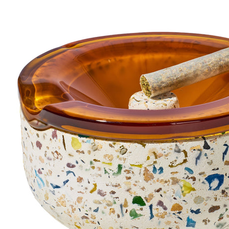 Symi Ashtray showcasing a terrazzo concrete base with amber glass insert, featuring a cork center and a pre-rolled cigarette resting in a groove.