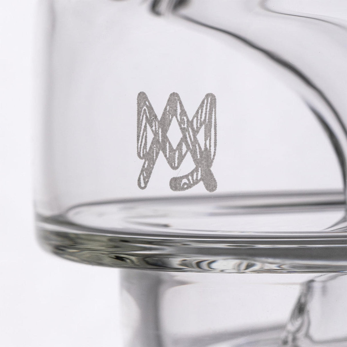 Close-up of Apex Dab Rig - Alpine Collection, showing a clear glass vessel with a silvery “MM” monogram etched on its side, highlighting its intricate design.