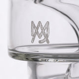 Close-up of Apex Dab Rig - Alpine Collection, showing a clear glass vessel with a silvery “MM” monogram etched on its side, highlighting its intricate design.