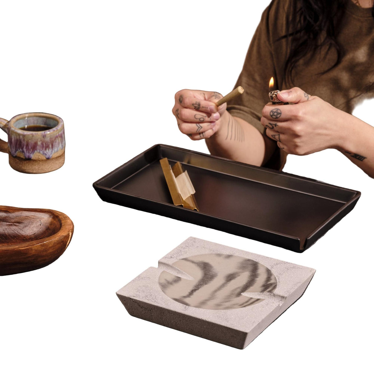 Tattooed hands roll a joint over the Roseland Tray Set, featuring a concrete ashtray, melamine tray, and a nearby ceramic mug and wooden bowl.