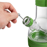 A close-up of a green and clear glass bong, with a hand holding a metal tool near the bowl. The product is shown against a white background