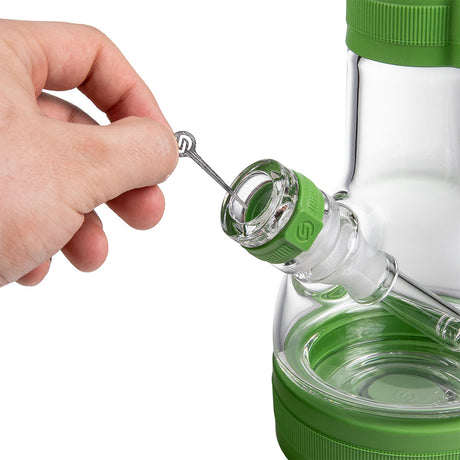 A close-up of a green and clear glass bong, with a hand holding a metal tool near the bowl. The product is shown against a white background