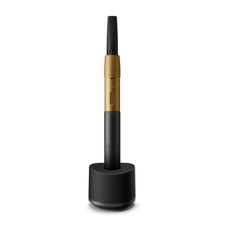 Base Charger - Black: Pen-style vaporizer with gold and black tones, seated upright in matching black charging dock, featuring a secure, weighted, non-slip design for Vessel devices.